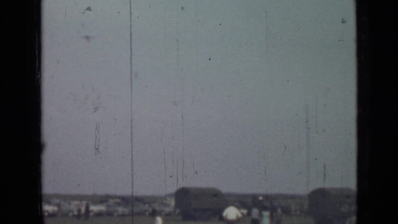 Pale sky and distant field seen through dark window bars, with cars and figures near the horizon.