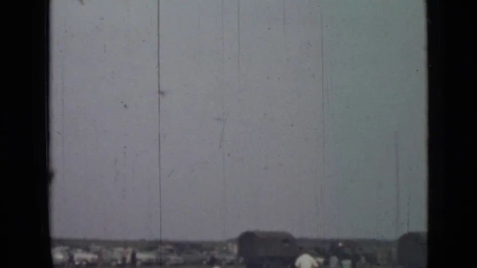 A 1967 8mm view across an open field in Saint Petersburg, with parked cars and small figures below a faded sky.