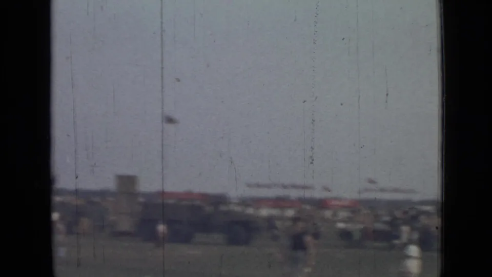 Distant 1967 home-movie view of a green field, low seating or parked forms, and scattered people rendered in soft focus.