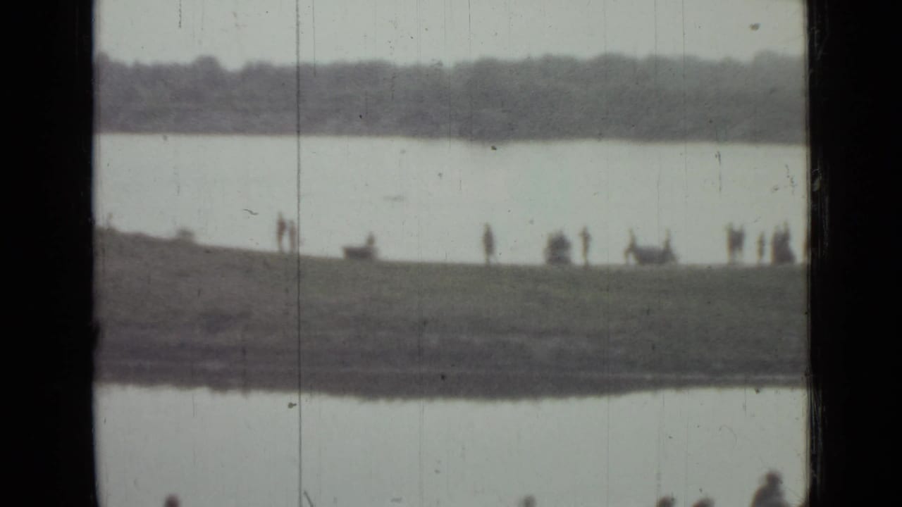 Distant 1967 Saint Petersburg riverbank scene with small groups of people on grass between water and a far tree line.