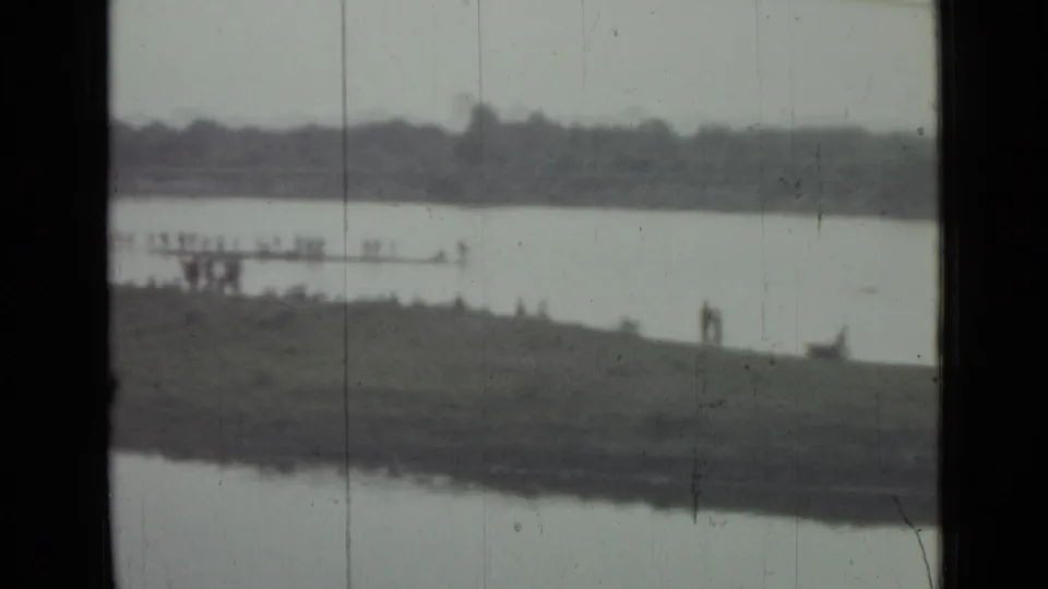 Wide home-movie frame of a Saint Petersburg riverside area with distant figures, water, grassy land, and a far shoreline.