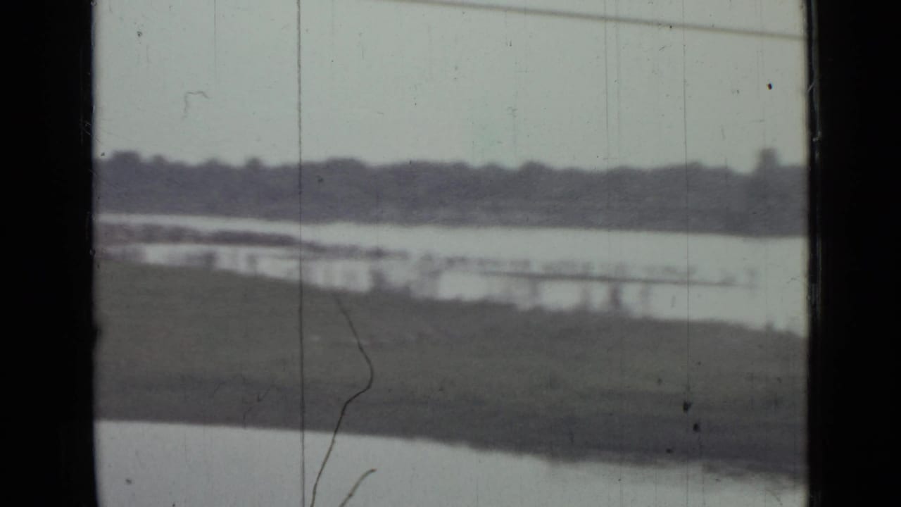 Soft 1967 8mm view near Saint Petersburg of a lake, grassy shore, distant treeline, low waterside structures, and dark window edges.