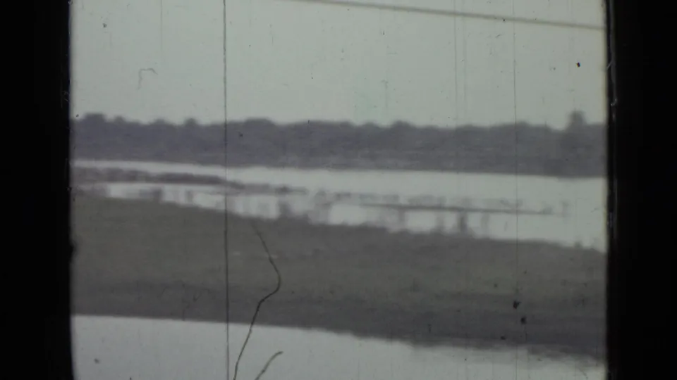 Soft 1967 8mm view near Saint Petersburg of a lake, grassy shore, distant treeline, low waterside structures, and dark window edges.