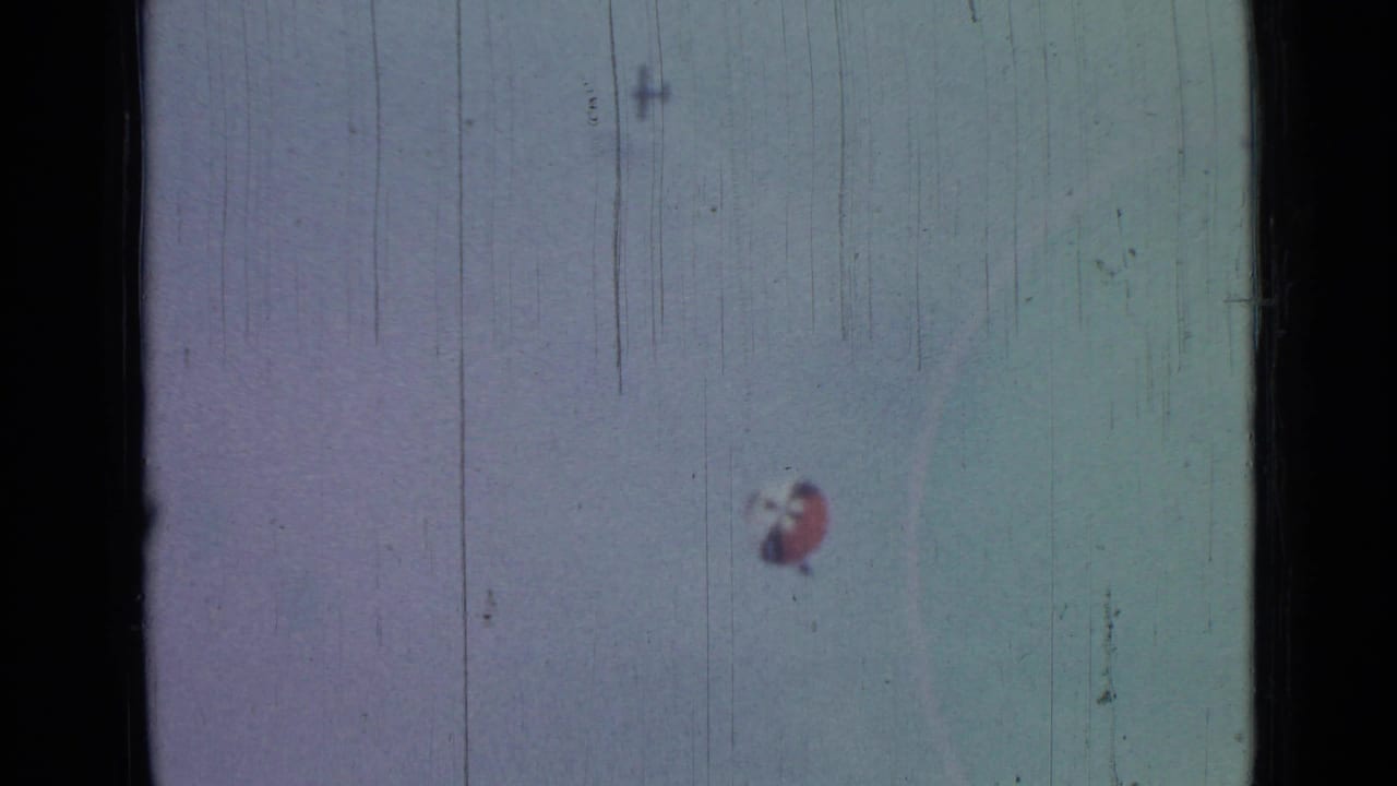 The distant parachute sits low in a pale sky, with 8mm grain, scratches, and dark borders visible across the frame.