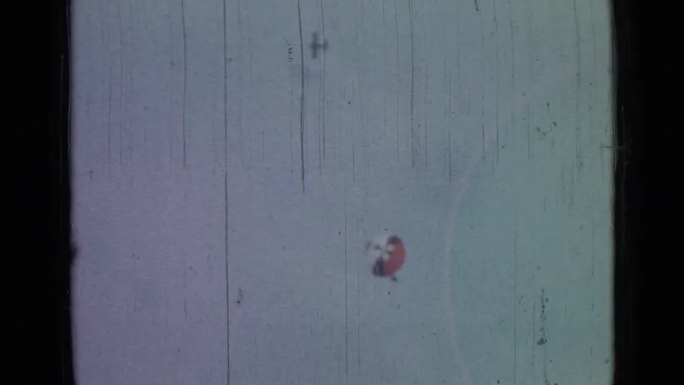 The distant parachute sits low in a pale sky, with 8mm grain, scratches, and dark borders visible across the frame.