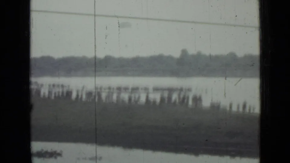 Distant spectators gather on a grassy waterfront in Saint Petersburg, Russia, with water and a tree-lined far shore visible through 8mm film wear.