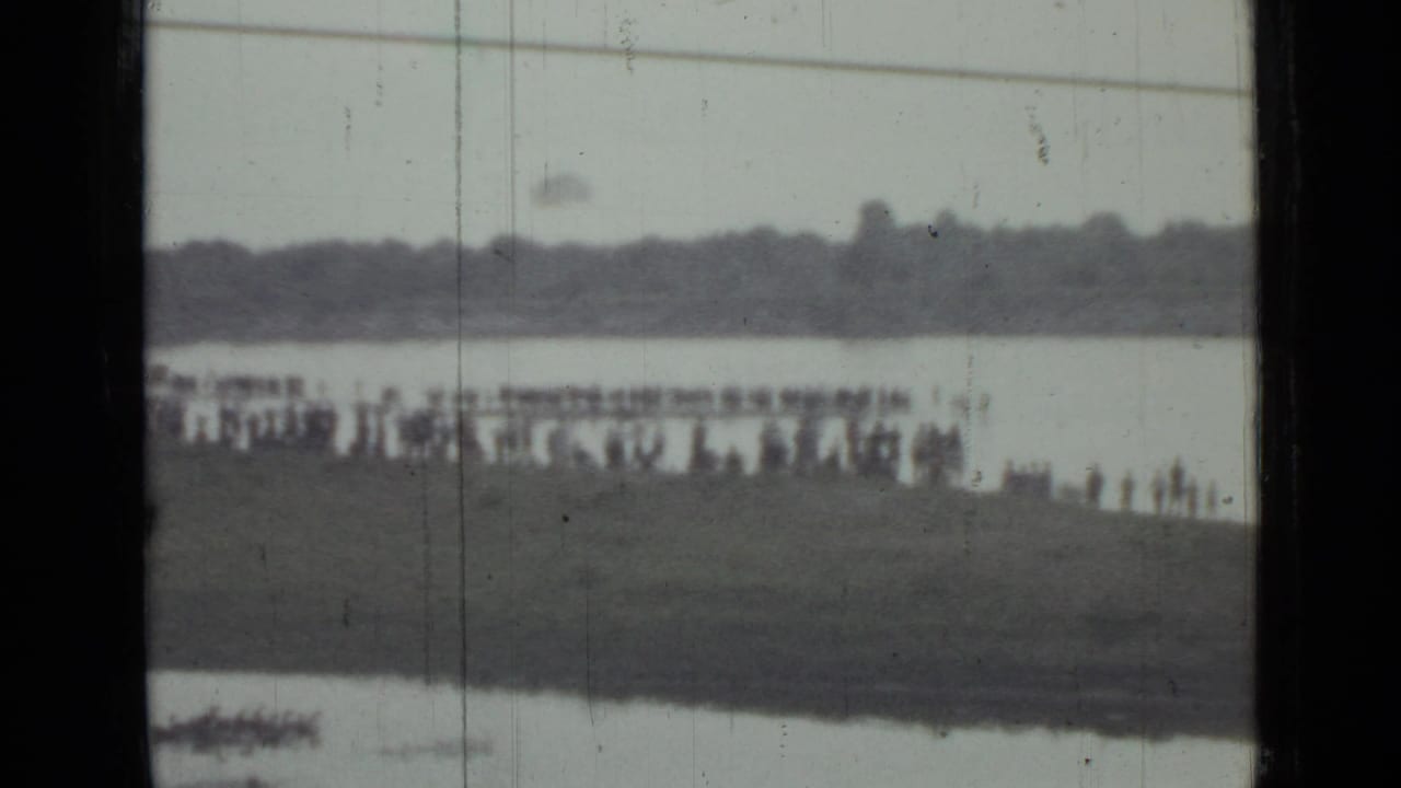 A distant 1967 Saint Petersburg crowd watches from a grassy bank near water, with the parachutist barely visible above the shoreline.