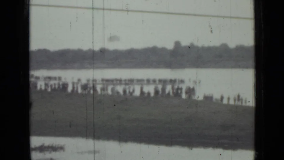 A distant 1967 Saint Petersburg crowd watches from a grassy bank near water, with the parachutist barely visible above the shoreline.