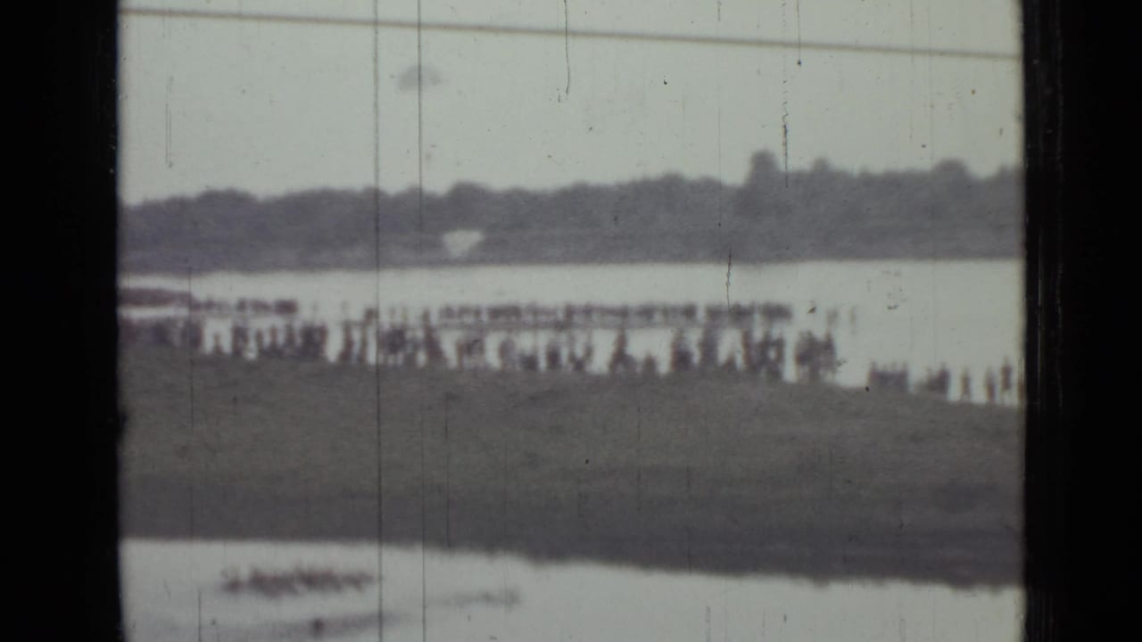 Distant crowd stands along a lakeside bank with a small boat on the water.