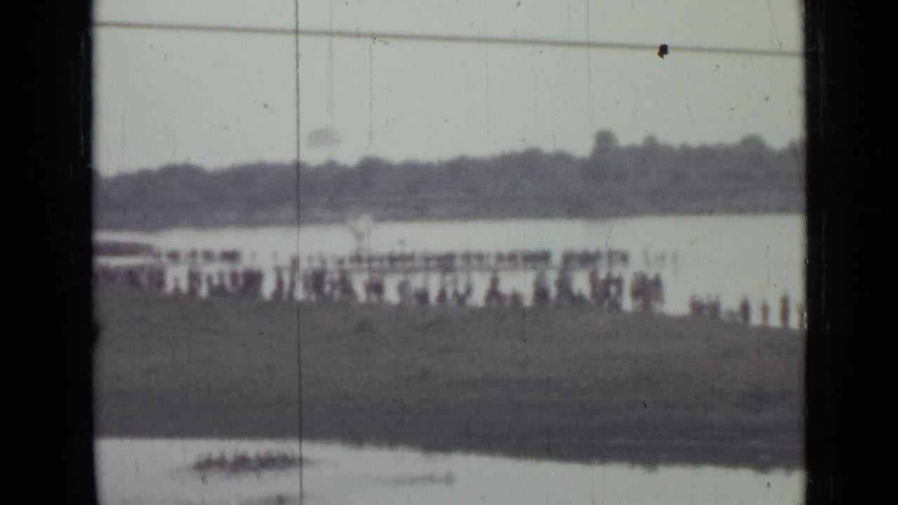 Soft 1967 Saint Petersburg lakeside frame with spectators along the bank, pale water behind them, and a distant wooded shoreline.