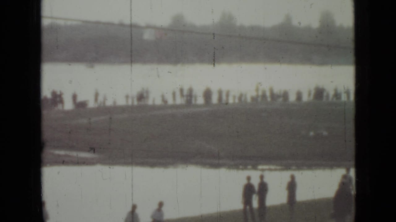 Distant people stand along a grassy shoreline beside a broad waterway under a gray sky.