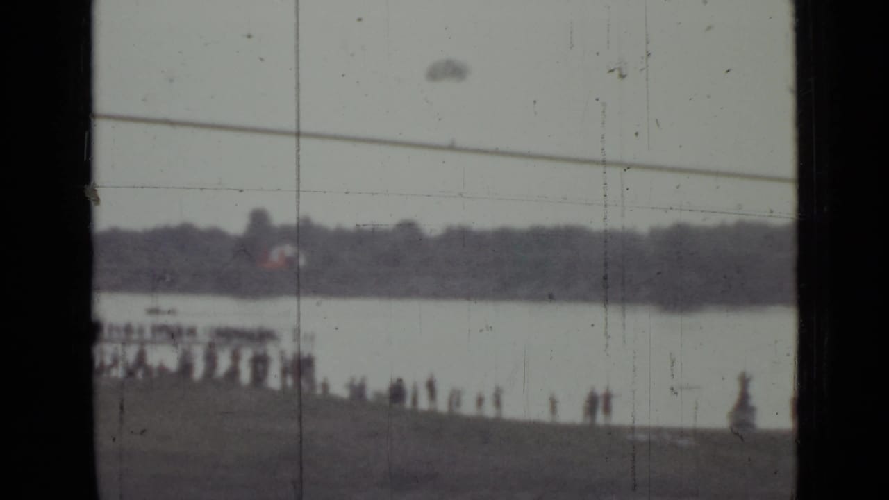 Soft 1967 home-movie frame of a Saint Petersburg shoreline crowd, waterway, small boat, and faint parachute-like object overhead.