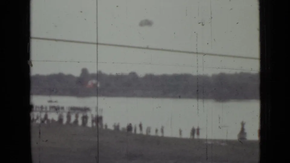 Soft 1967 home-movie frame of a Saint Petersburg shoreline crowd, waterway, small boat, and faint parachute-like object overhead.