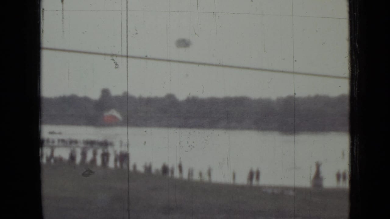 Distant riverbank crowd watches a parachute descending over water beneath a utility line.
