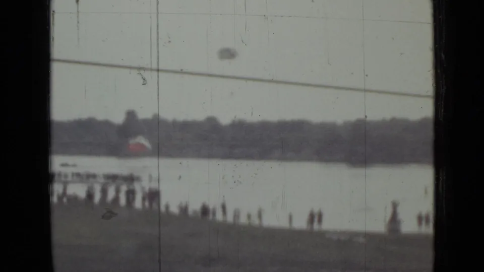 1967 Saint Petersburg home-movie frame showing a distant crowd on a riverbank as a parachute descends over the water.
