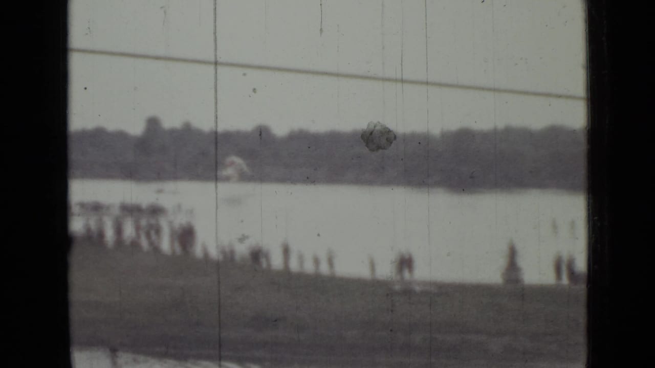 Distant lakeshore with spectators and a treeline seen through scratched 8mm film.