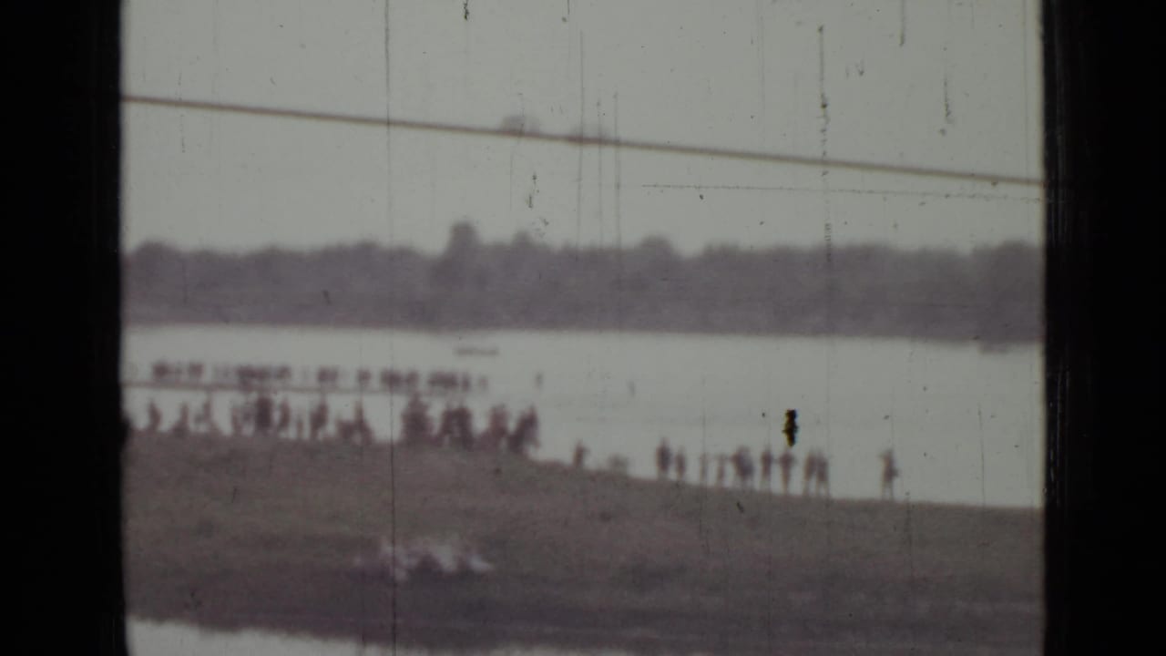 1967 Saint Petersburg home-movie frame showing small figures gathered on a shoreline across a wide, pale water surface.
