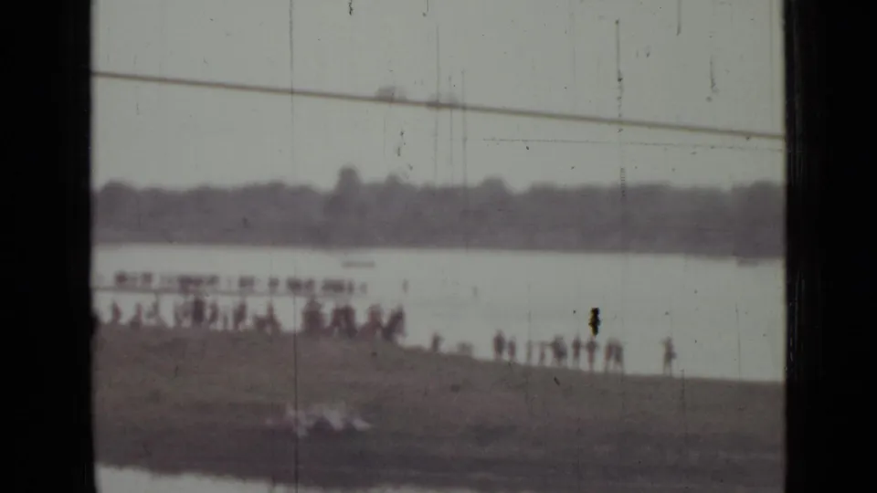1967 Saint Petersburg home-movie frame showing small figures gathered on a shoreline across a wide, pale water surface.