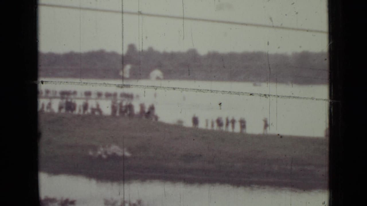 Distant spectators stand along a riverbank with water and treeline behind them.