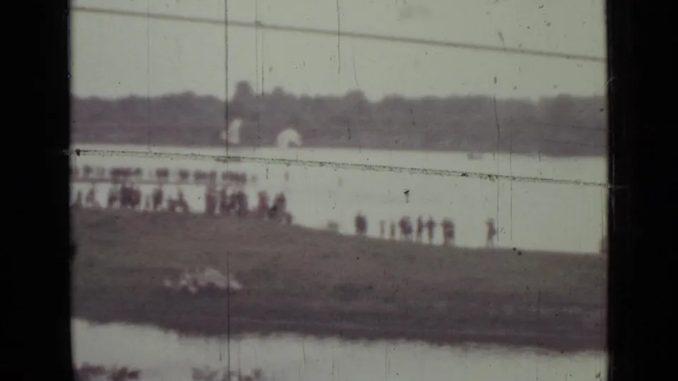 1967 Saint Petersburg home-movie frame showing distant people gathered on a grassy riverbank beside broad water and a hazy treeline.