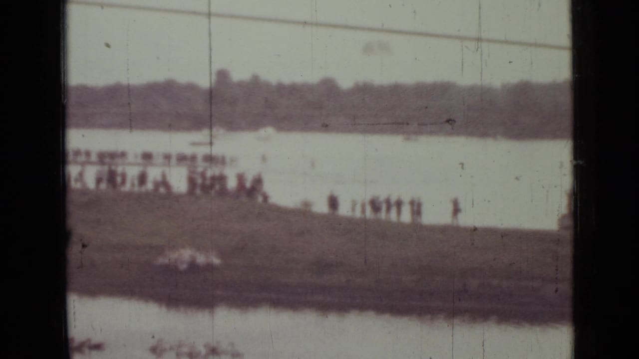 Distant 8mm view of spectators on a Saint Petersburg riverbank as a pale parachute shape appears across the water.