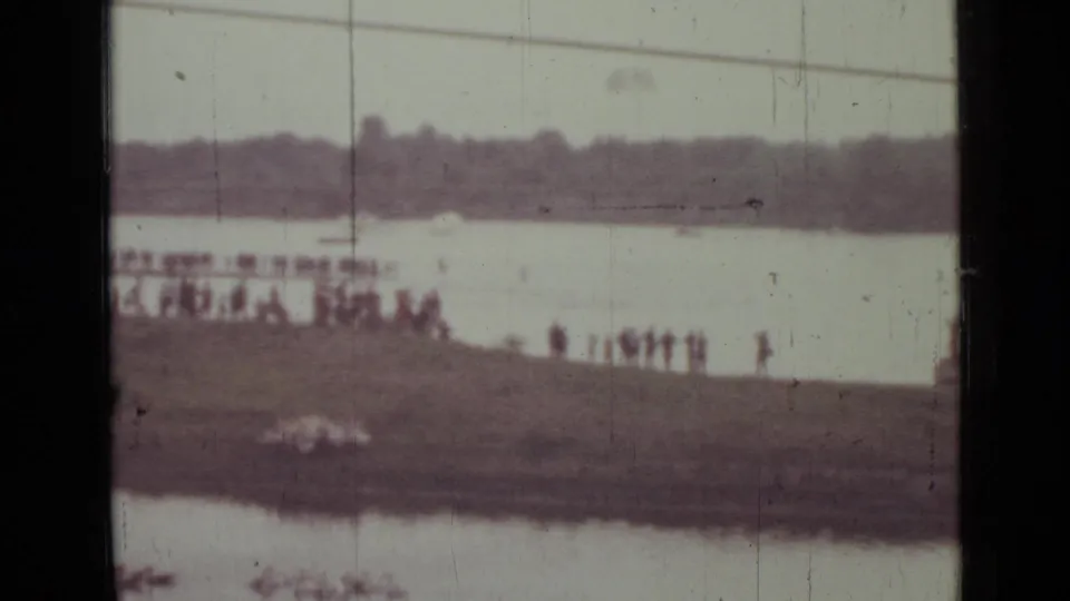 Distant 8mm view of spectators on a Saint Petersburg riverbank as a pale parachute shape appears across the water.