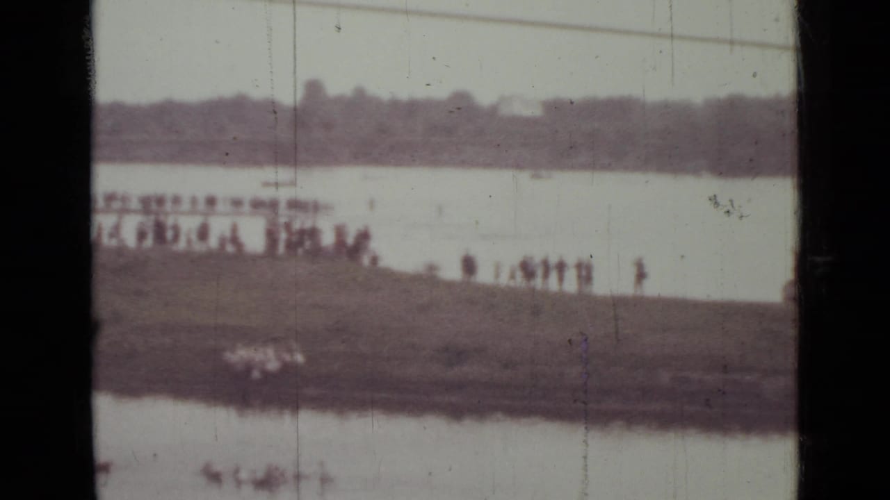 Soft, faded 1967 home-movie frame of people watching from a grassy Saint Petersburg riverbank, with water and trees beyond.