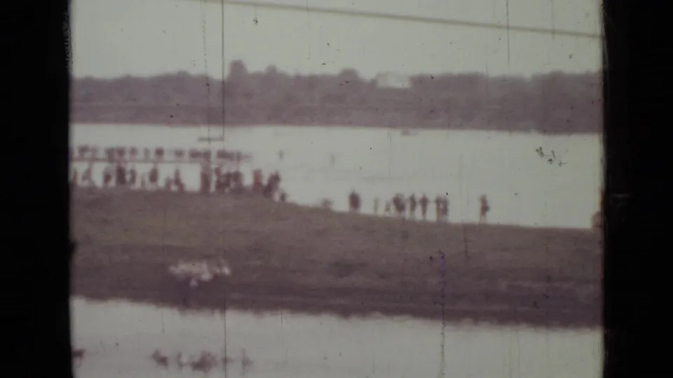 Soft, faded 1967 home-movie frame of people watching from a grassy Saint Petersburg riverbank, with water and trees beyond.