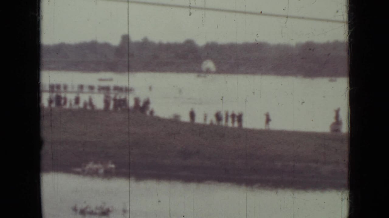 People stand along a distant pier beside calm water with shoreline beyond.