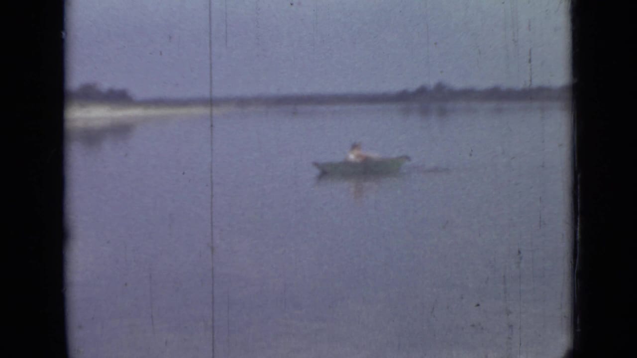 Distant man in a small canoe on calm water near a pale shoreline.