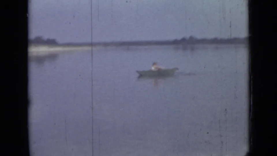 1967 8mm frame of a distant man canoeing alone on calm water, with a low shoreline and trees in the background.