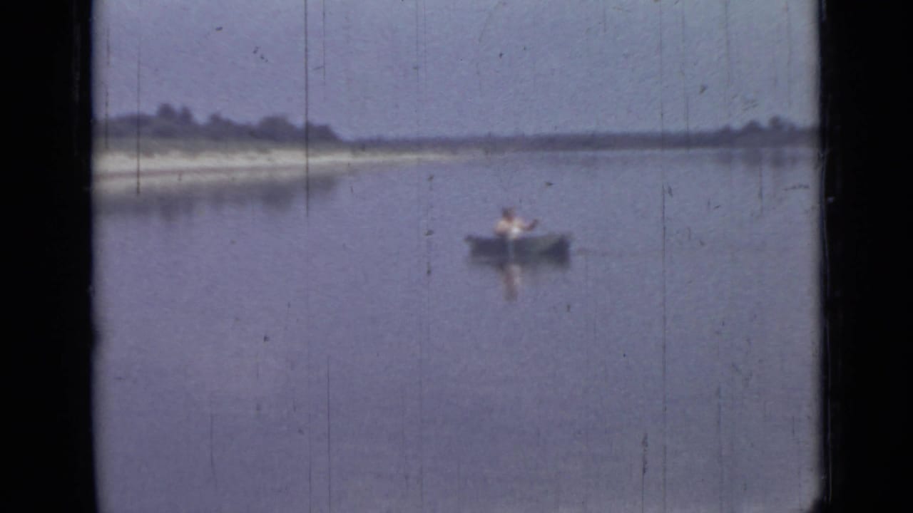 The 8mm home-movie frame shows a lone man in a canoe on quiet water, with muted color and a low wooded shoreline.