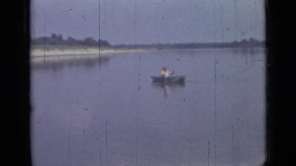 The 8mm home-movie frame shows a lone man in a canoe on quiet water, with muted color and a low wooded shoreline.