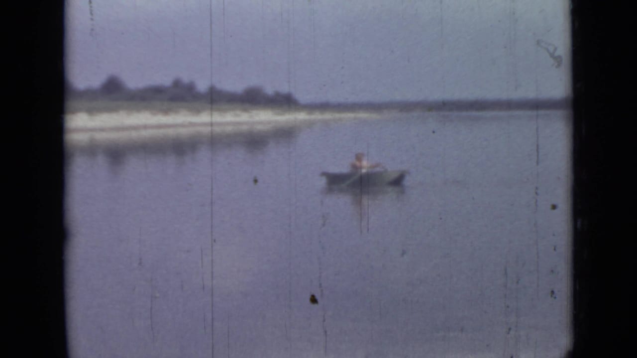 Final 1967 8mm frame of a lone canoeist on blue-gray water, with shoreline, trees and visible film wear.