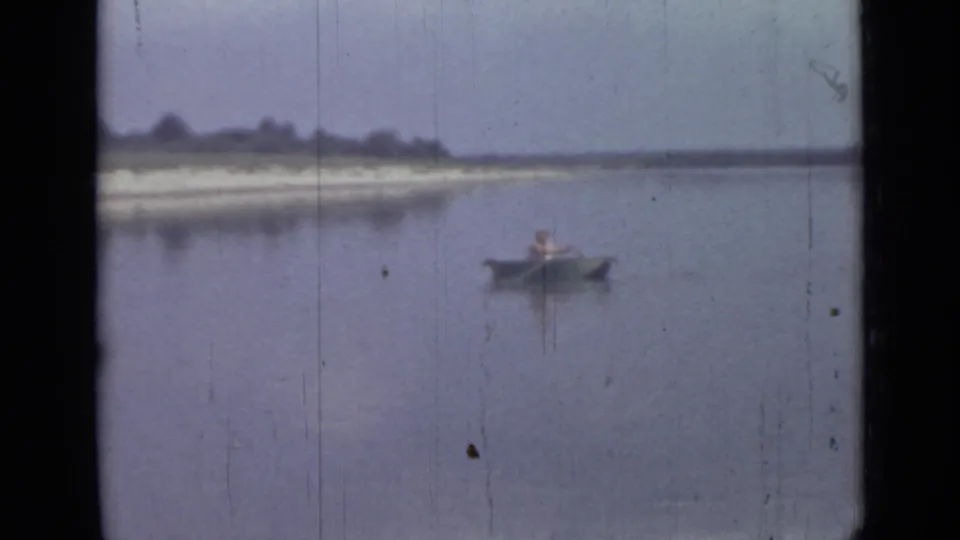 Final 1967 8mm frame of a lone canoeist on blue-gray water, with shoreline, trees and visible film wear.