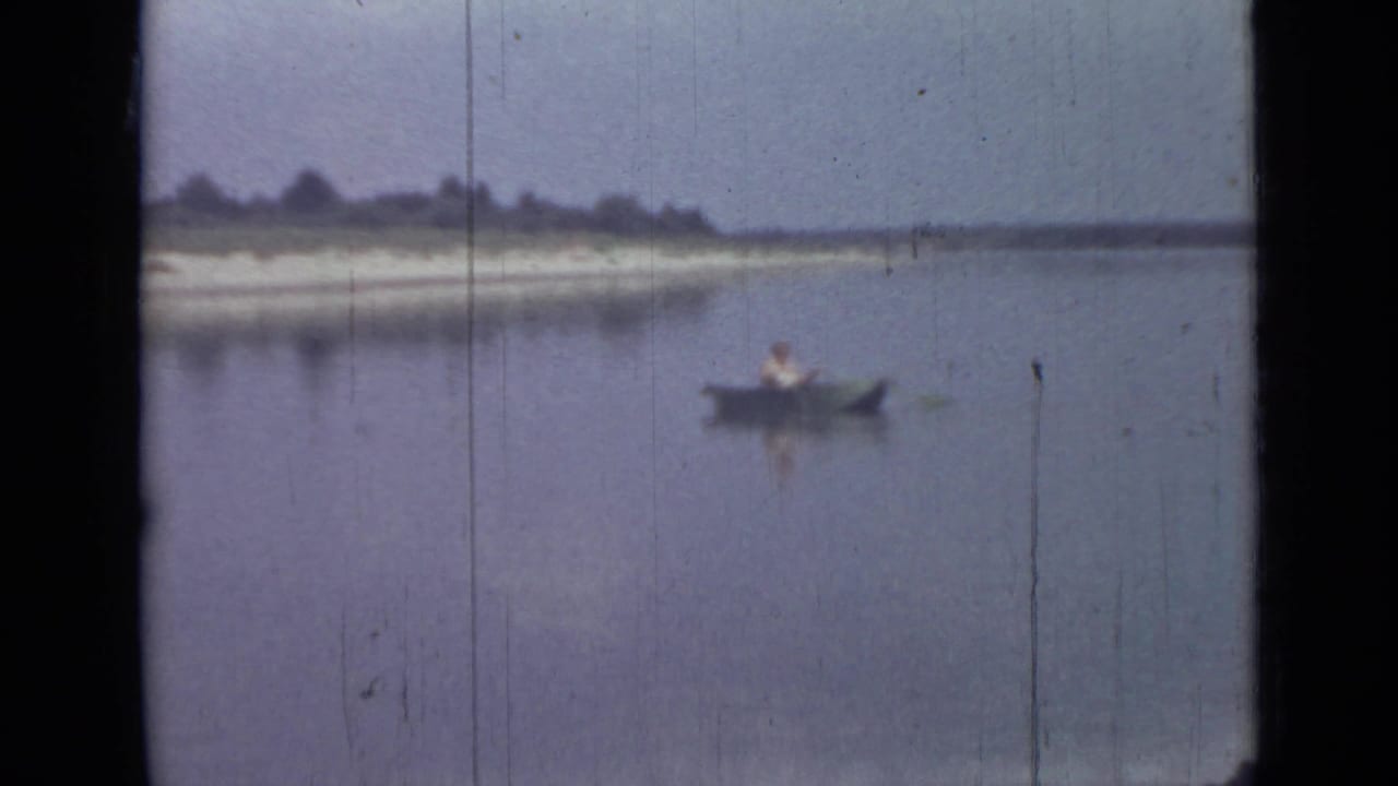 1967 8mm home-movie frame from Saint Petersburg showing one person rowing a small boat across a calm lake near a distant shore.