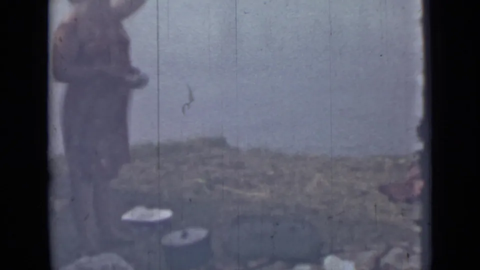 Soft 1967 8mm frame from Saint Petersburg showing a woman by a hazy riverbank with picnic or food-preparation items nearby.