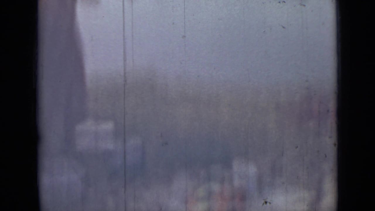 The frame catches part of the woman at the river edge, with indistinct supplies or foreground objects softened by blur and film wear.