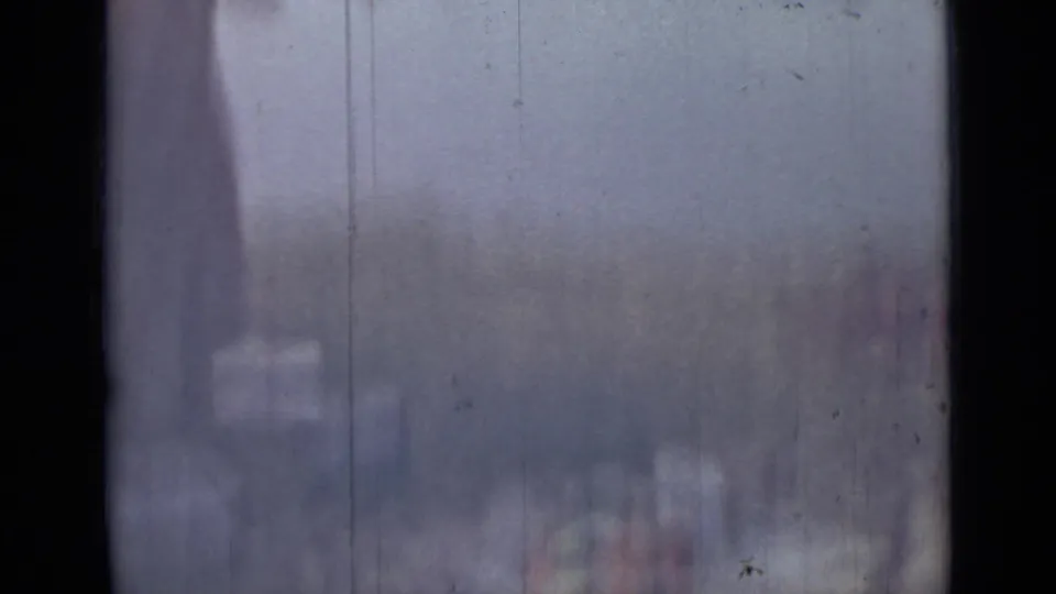 The frame catches part of the woman at the river edge, with indistinct supplies or foreground objects softened by blur and film wear.
