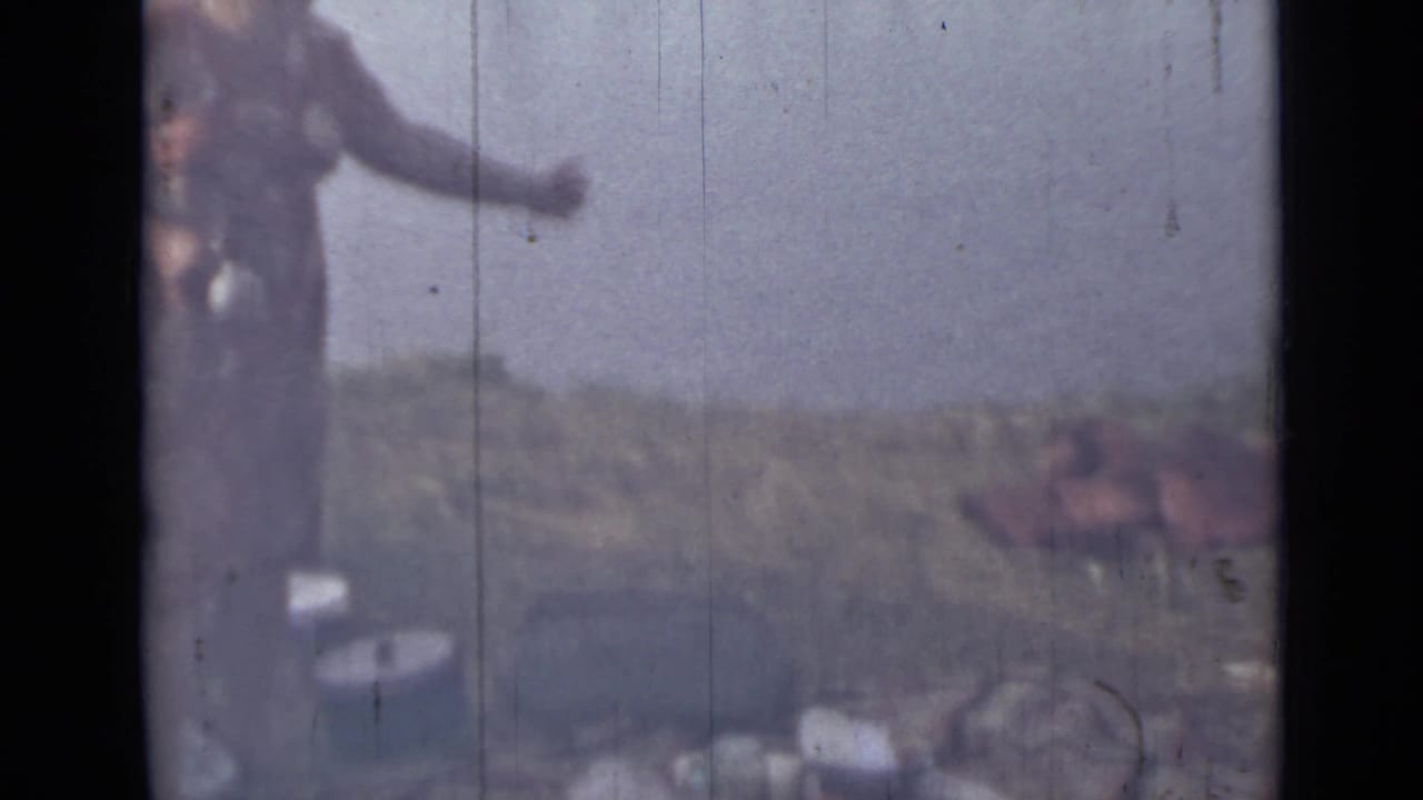 A 1967 Saint Petersburg home-movie frame shows the woman extending an arm near riverside containers, with water and grassy bank behind her.