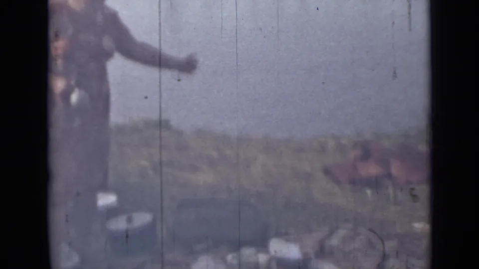 A 1967 Saint Petersburg home-movie frame shows the woman extending an arm near riverside containers, with water and grassy bank behind her.