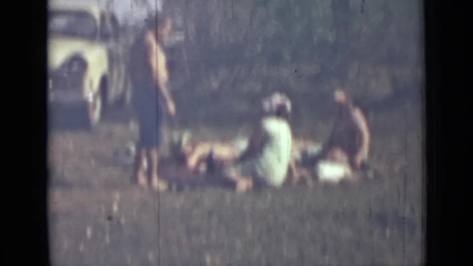 1967 8mm home-movie frame of people picnicking or sunbathing on grass near Saint Petersburg, with a parked car in the background.