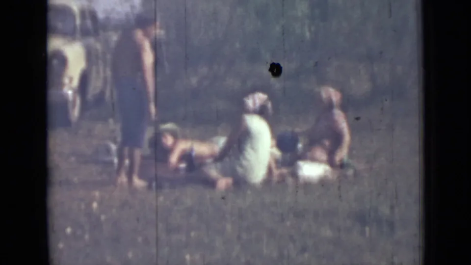Home-movie view of a small outdoor group relaxing on grass in Saint Petersburg, Russia, with a vintage car partly visible behind them.