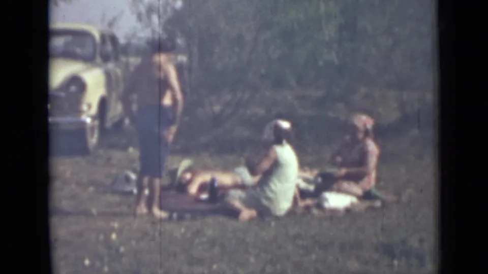 1967 8mm frame showing a group in swimwear or summer clothing gathered on grass near a light-colored car and shrubs.