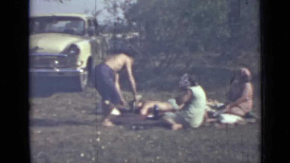 Saint Petersburg 1967 home-movie frame of a man bending near a relaxed picnic group, with a light-colored car and trees behind them.