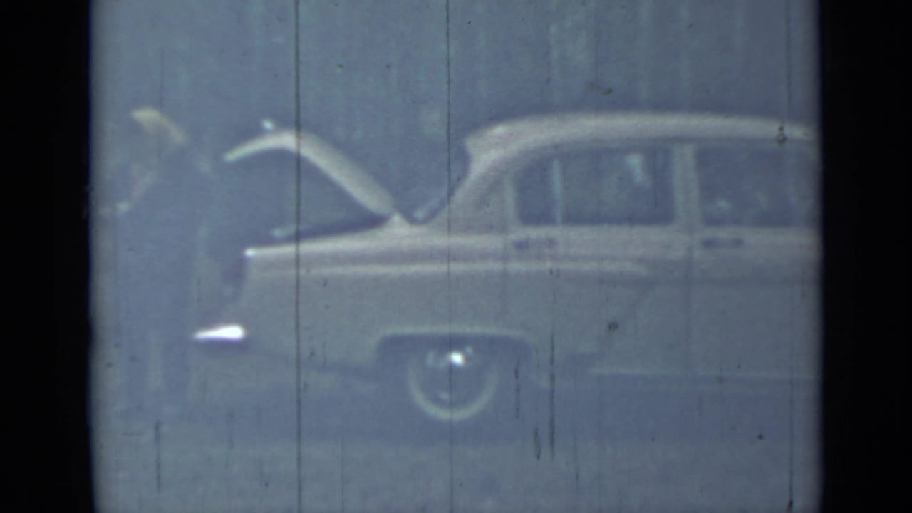 Person beside a pale sedan with its trunk open in a wooded outdoor area.