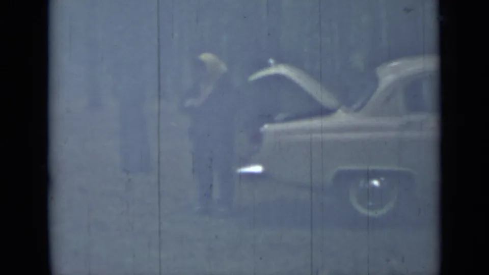 The vintage sedan remains framed with its trunk open, while a person stands nearby in soft, scratched 8mm footage.
