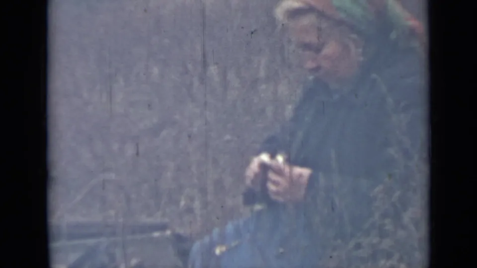 A bundled woman in a headscarf works close to the ground with a potato and knife, with a bucket partly visible nearby.