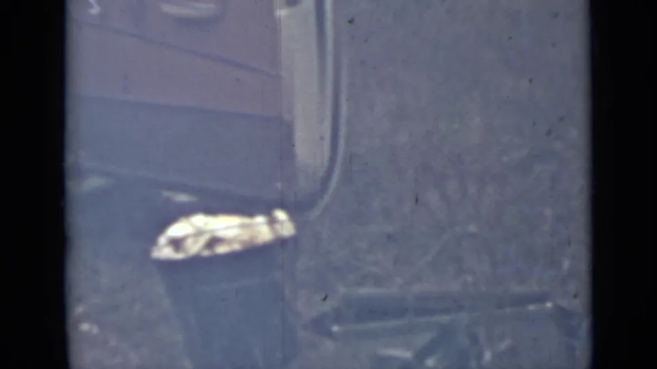 The frame shifts toward a partial vehicle or outdoor object while the bundled woman remains near the edge of the home-movie frame.