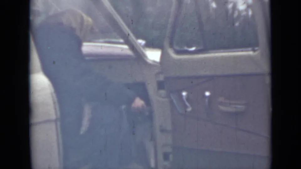 In the 1967 Saint Petersburg home-movie frame, a woman in a dark coat and light head covering leans into an open vehicle.